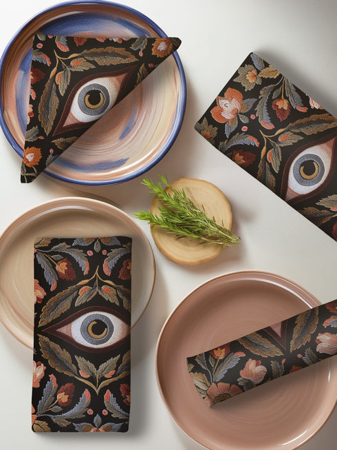 Set of four square black polyester cloth napkins featuring a central embroidered-style eye motif framed by floral and leaf patterns in autumnal tones of rust, ochre, and gold. The design is printed on one side, with a white reverse. Each napkin measures 18x18 inches.